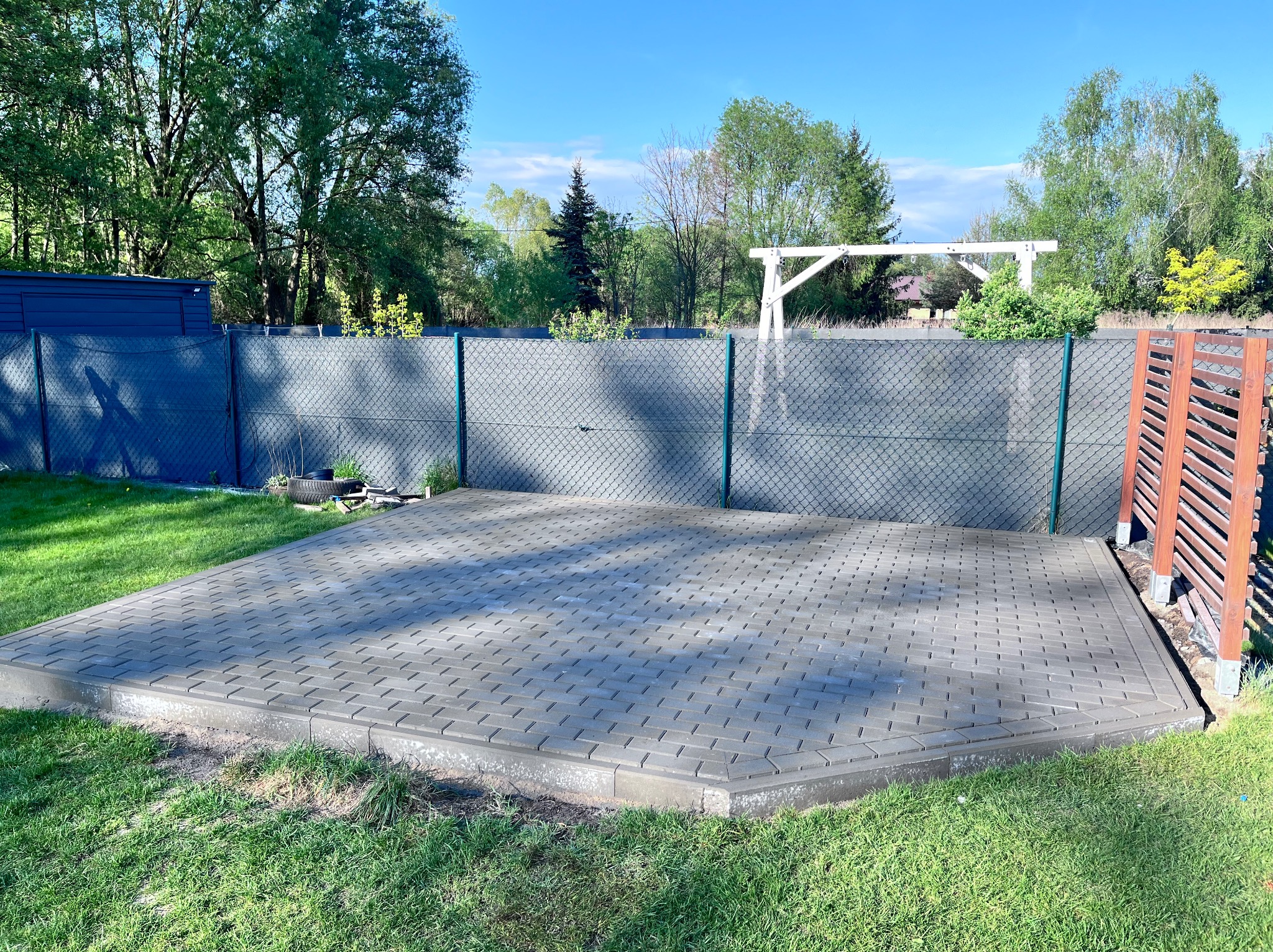 Nowo ułożona, szara kostka brukowa tworząca taras w ogrodzie w Poznaniu, otoczona świeżą zielenią trawnika i ogrodzeniem z siatki.