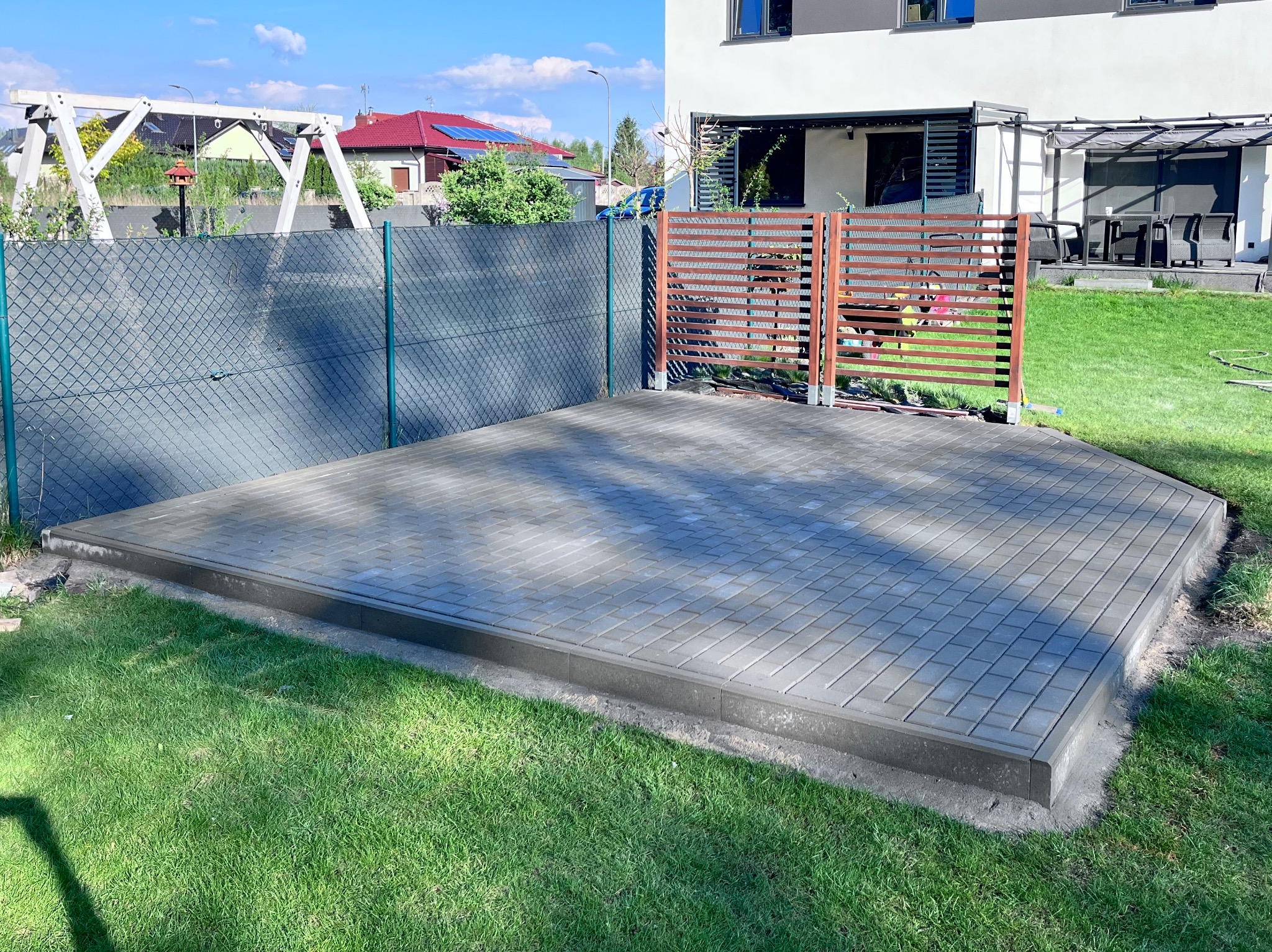 Wyrównana powierzchnia z szarej kostki brukowej na trawniku, obrzeża wykończone betonową ramą, w tle ogrodzenie panelowe i drewniane, dom z jasną elewacją.