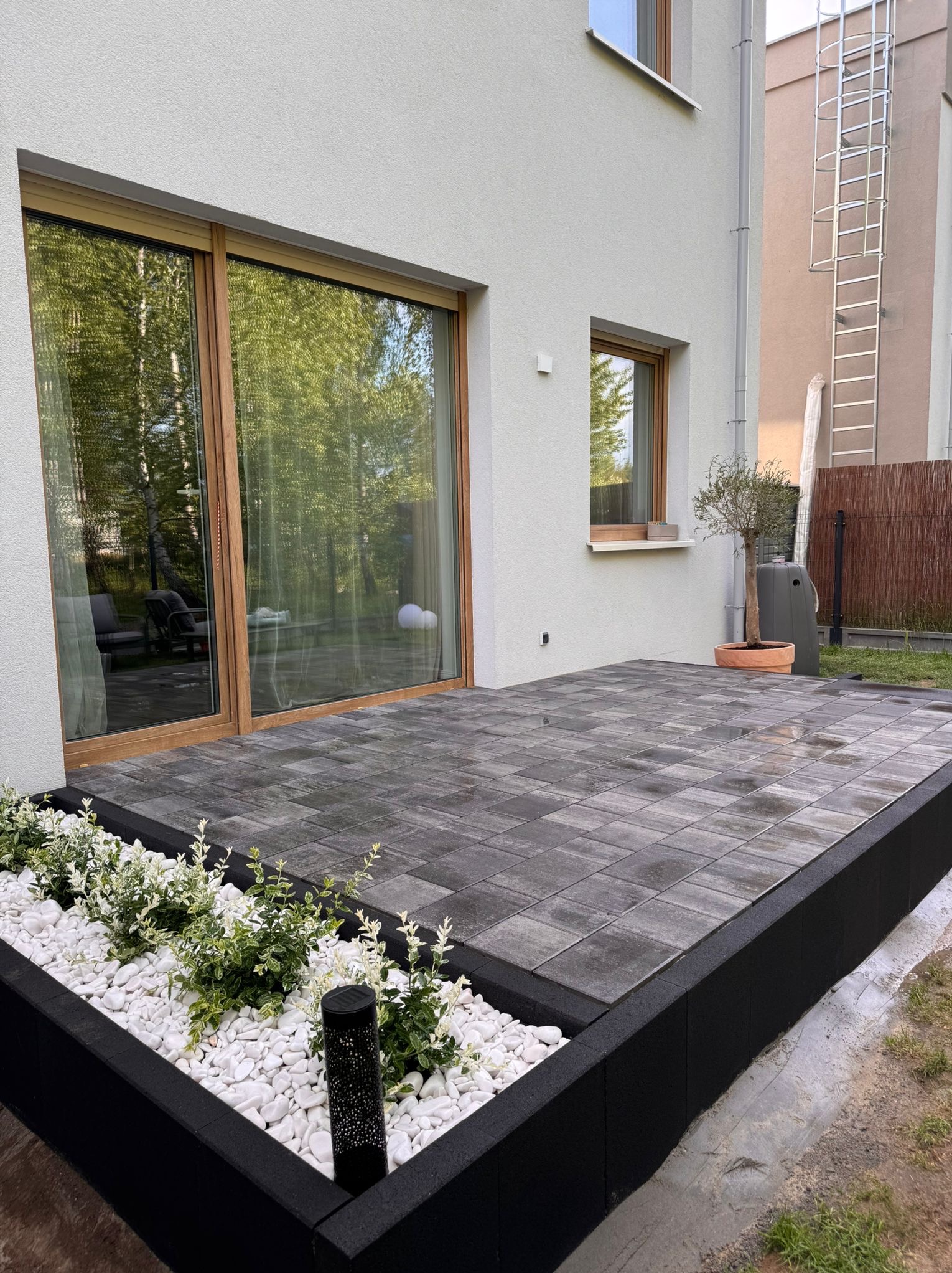 Nowoczesny taras z szarej kostki brukowej, czarna obwódka, białe kamyki i rośliny. Duże okno tarasowe z drewnianą ramą. Minimalistyczny design.