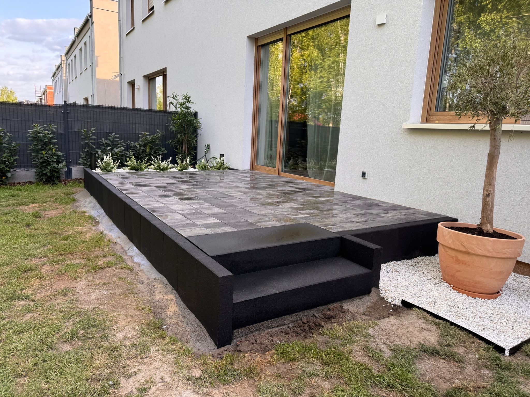 Nowoczesny taras z szarej kostki brukowej i czarnymi schodami, z donicą z drzewkiem oliwnym na białym żwirze. W tle dom z dużymi oknami i grafitowe ogrodzenie.