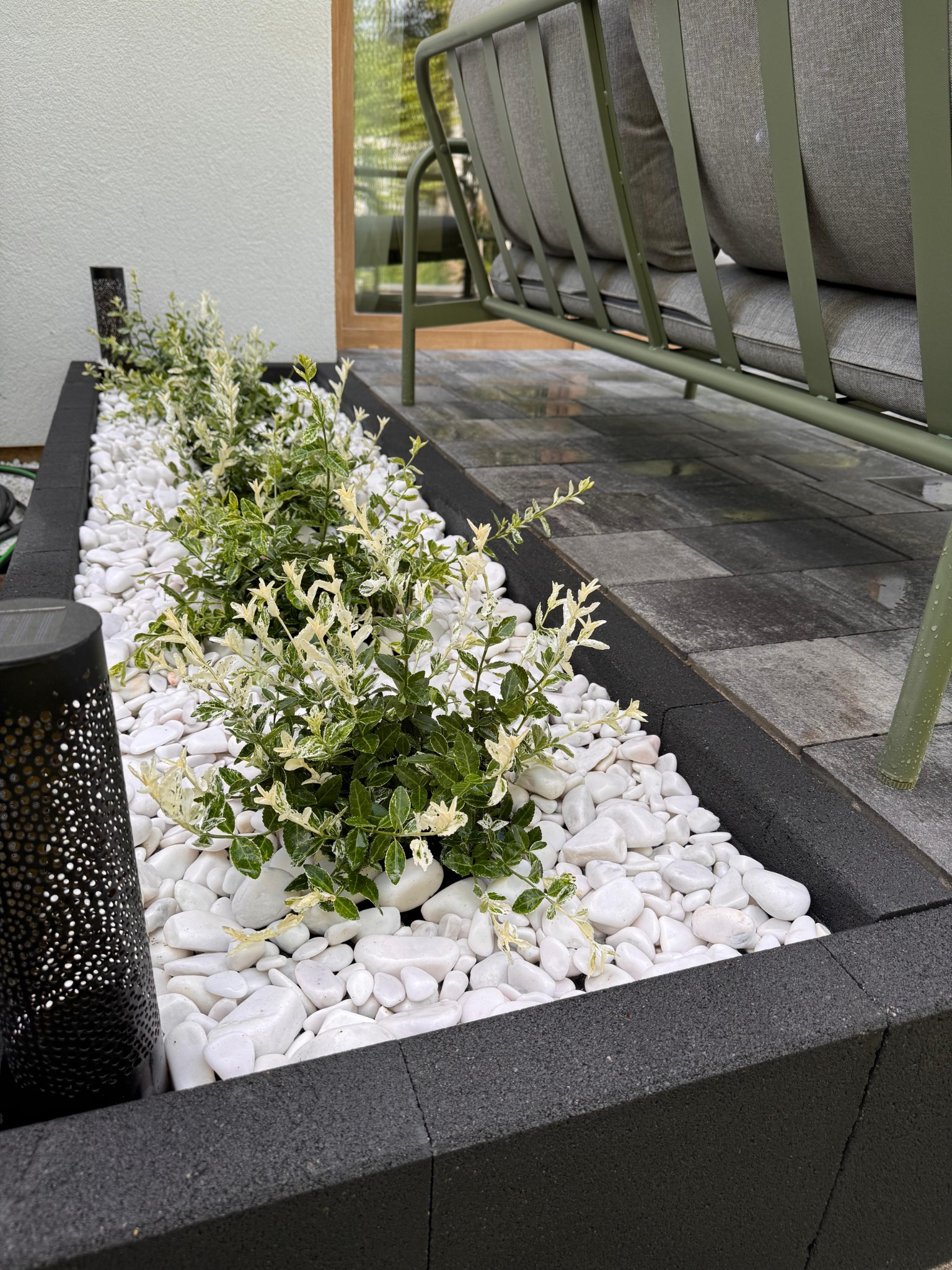 Obrzeże rabaty z czarnych kostek, wypełnione białymi kamieniami i zielonymi krzewami. W tle fragment szarej kostki brukowej i zielony metalowy mebel ogrodowy.