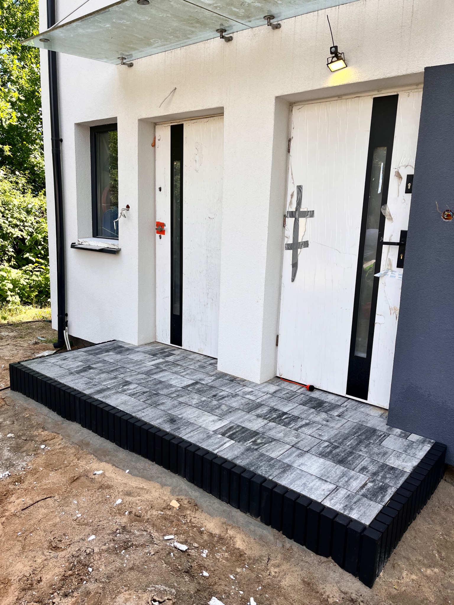 Nowy podest wejściowy z szarej kostki brukowej z czarnym obrzeżem przed białym budynkiem, widok z dołu. Prace brukarskie w toku, niezakończone.