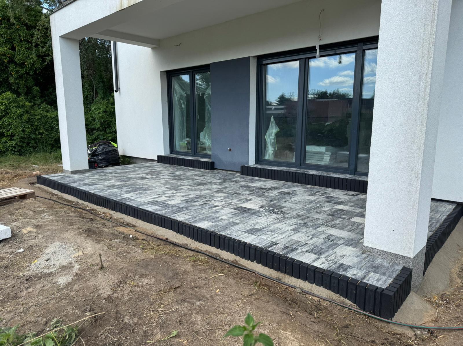 Nowoczesny taras z szarej kostki brukowej obramowany czarnym obrzeżem, przylegający do budynku z dużymi oknami. Widoczne elementy wykończeniowe.