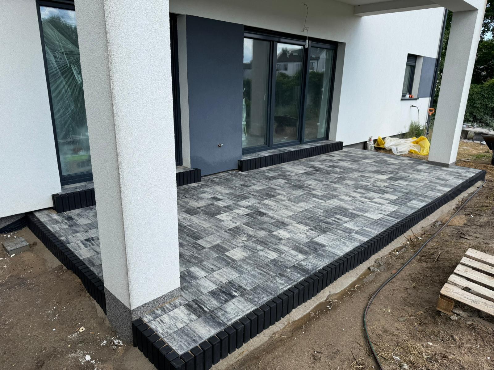 Wykończony taras z szarej kostki brukowej, obrzeża z czarnych elementów. Widok na dom z dużymi oknami. Częściowo ułożona kostka, widoczne przygotowania do dalszych prac.