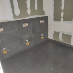 Ciemnoszare płytki podłogowe i ścienne w trakcie montażu w łazience, widoczne przygotowane punkty instalacji wodnej i fragmenty płyt kartonowo-gipsowych na ścianach przed szpachlowaniem.