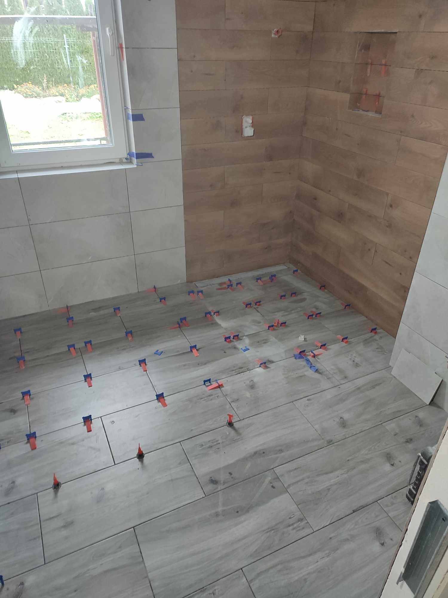 Układanie płytek imitujących drewno na podłodze i ścianach w łazience, z użyciem klipsów poziomujących i taśmy malarskiej przy oknie. Widoczna wnęka w ścianie wyłożonej płytkami drewnopodobnymi.