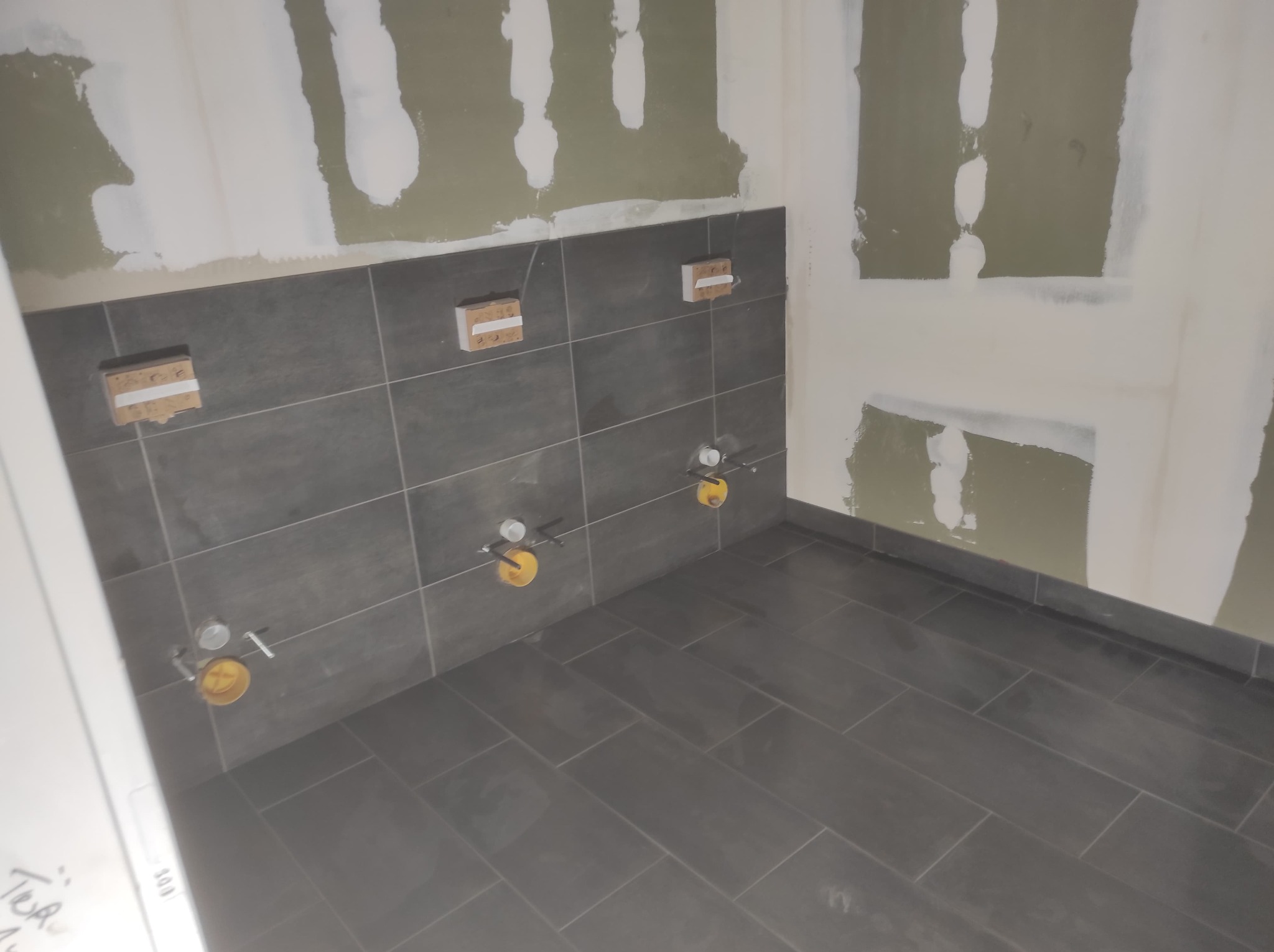 Ciemnoszare płytki podłogowe i ścienne w trakcie montażu w łazience, widoczne przygotowane punkty instalacji wodnej i fragmenty płyt kartonowo-gipsowych na ścianach przed szpachlowaniem.