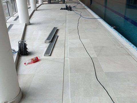 Jasne płytki podłogowe wokół basenu, z widocznymi kratkami odpływowymi, zwiniętym kablem, butelką po napoju i sprzętem do czyszczenia w tle.