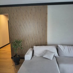 Grzegorz Podleszuk - Nowoczesny salon z drewnianą ścianką działową z lameli, szarą sofą i rośliną w czarnej donicy. Parkiet na podłodze, minimalistyczny design.