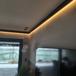 Grzegorz Podleszuk - Nowoczesne wnętrze z oświetleniem LED wzdłuż sufitu, wiszące lampy kuliste i widok na zewnątrz przez okno. Minimalistyczny design z roślinami doniczkowymi.