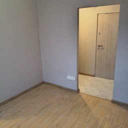 Grzegorz Podleszuk - Jasne panele podłogowe imitujące drewno w nowym pokoju z szarą ścianą i widokiem na przedpokój z beżową podłogą i drzwiami.