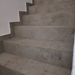 Grzegorz Podleszuk - Schody wykończone szarą, kamienną okładziną, widok z dołu. Minimalistyczny design, jasne ściany, precyzyjne wykończenie krawędzi. Detal architektoniczny.