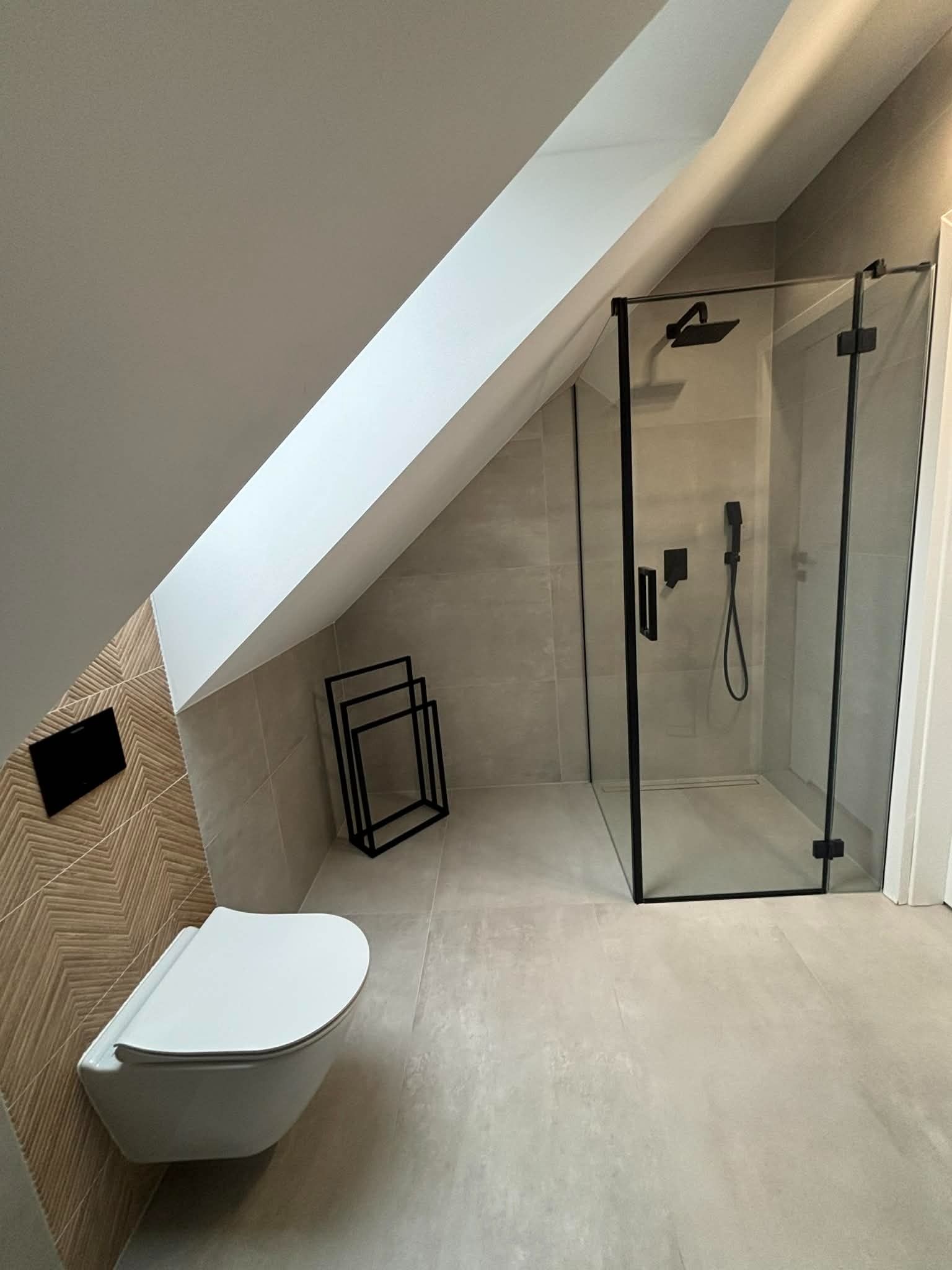 Nowoczesna łazienka na poddaszu ze skośnym sufitem. Kabina prysznicowa z czarnymi ramami, podwieszana toaleta i dekoracyjna ściana z wzorem jodełki.