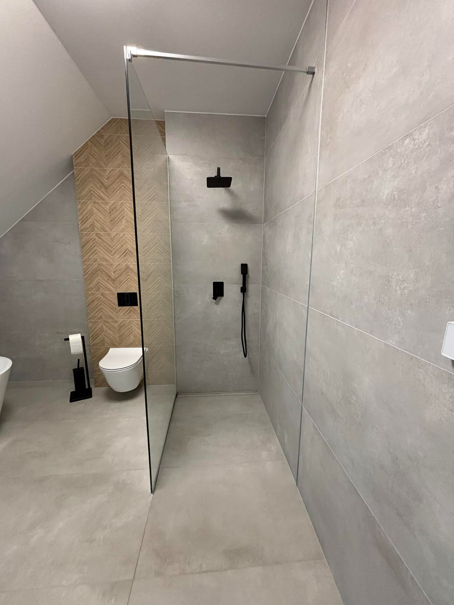 Nowoczesna łazienka w Gorzowie Wielkopolskim z szarą glazurą, kabiną typu walk-in i toaletą. Uwagę zwraca ściana z drewnopodobną mozaiką w jodełkę.