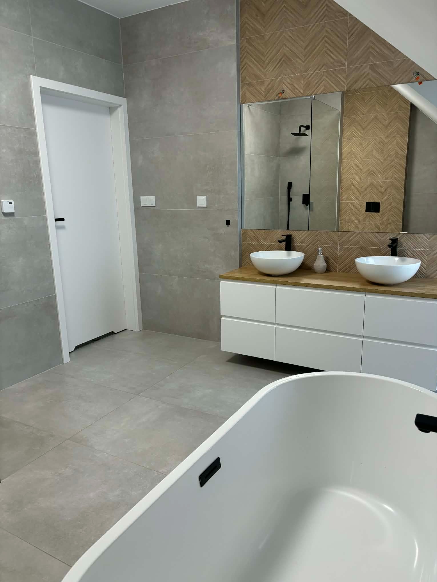 Nowoczesna łazienka na poddaszu z szarą płytką, umywalkami nablatowymi i drewnopodobną mozaiką nad lustrem. Biała wanna wolnostojąca na pierwszym planie.