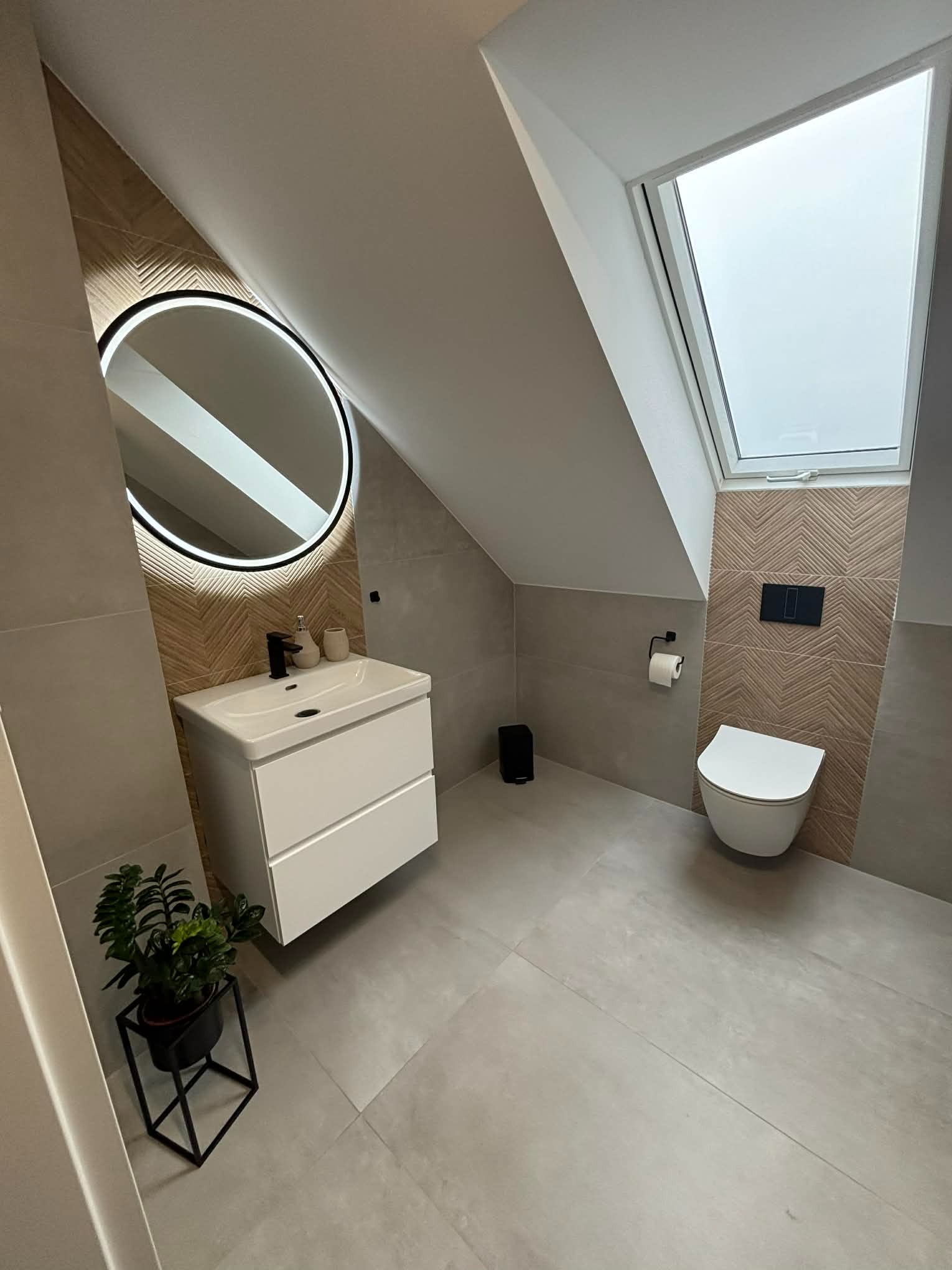 Nowoczesna łazienka na poddaszu z oknem dachowym, umywalką z szafką, okrągłym lustrem z podświetleniem LED i toaletą. Na ścianach płytki w odcieniach szarości i beżu.