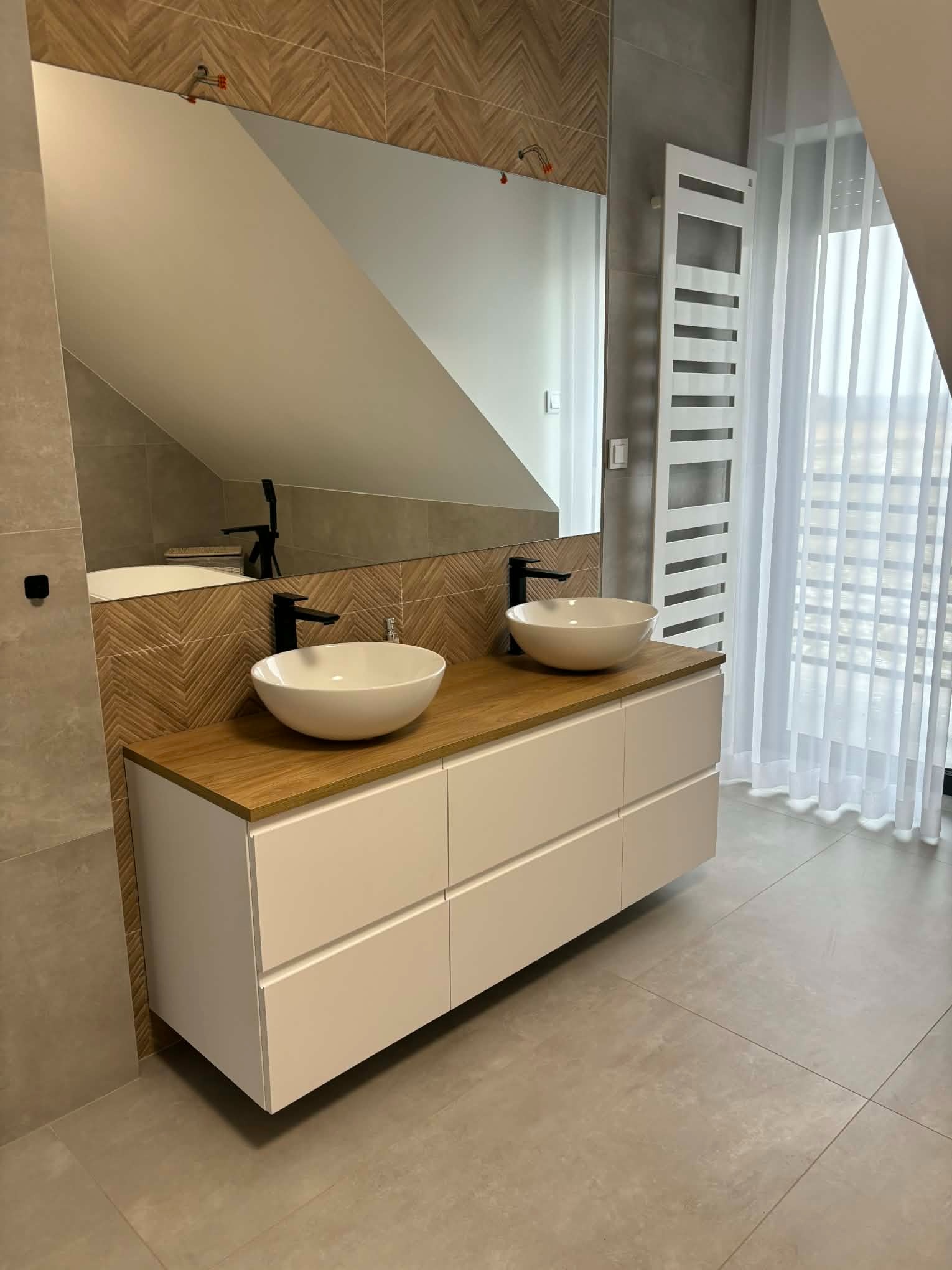 Nowoczesna łazienka na poddaszu: biała szafka z drewnianym blatem, dwie umywalki nablatowe, lustro, grzejnik drabinkowy i okno z firanką.