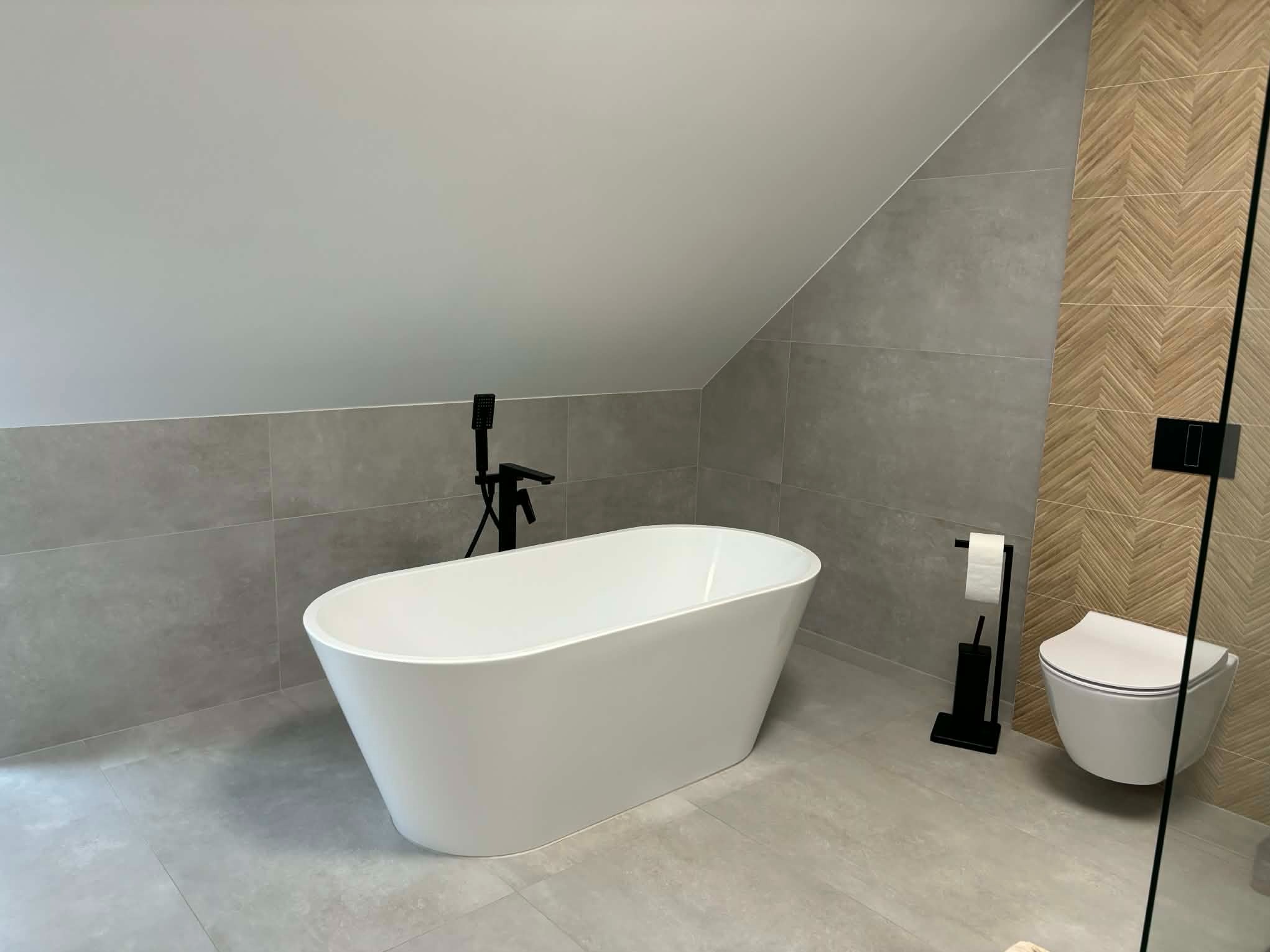 Nowoczesna łazienka na poddaszu z wolnostojącą białą wanną i toaletą w stylu minimalistycznym. Szare płytki i drewnopodobne akcenty. Czarna armatura.