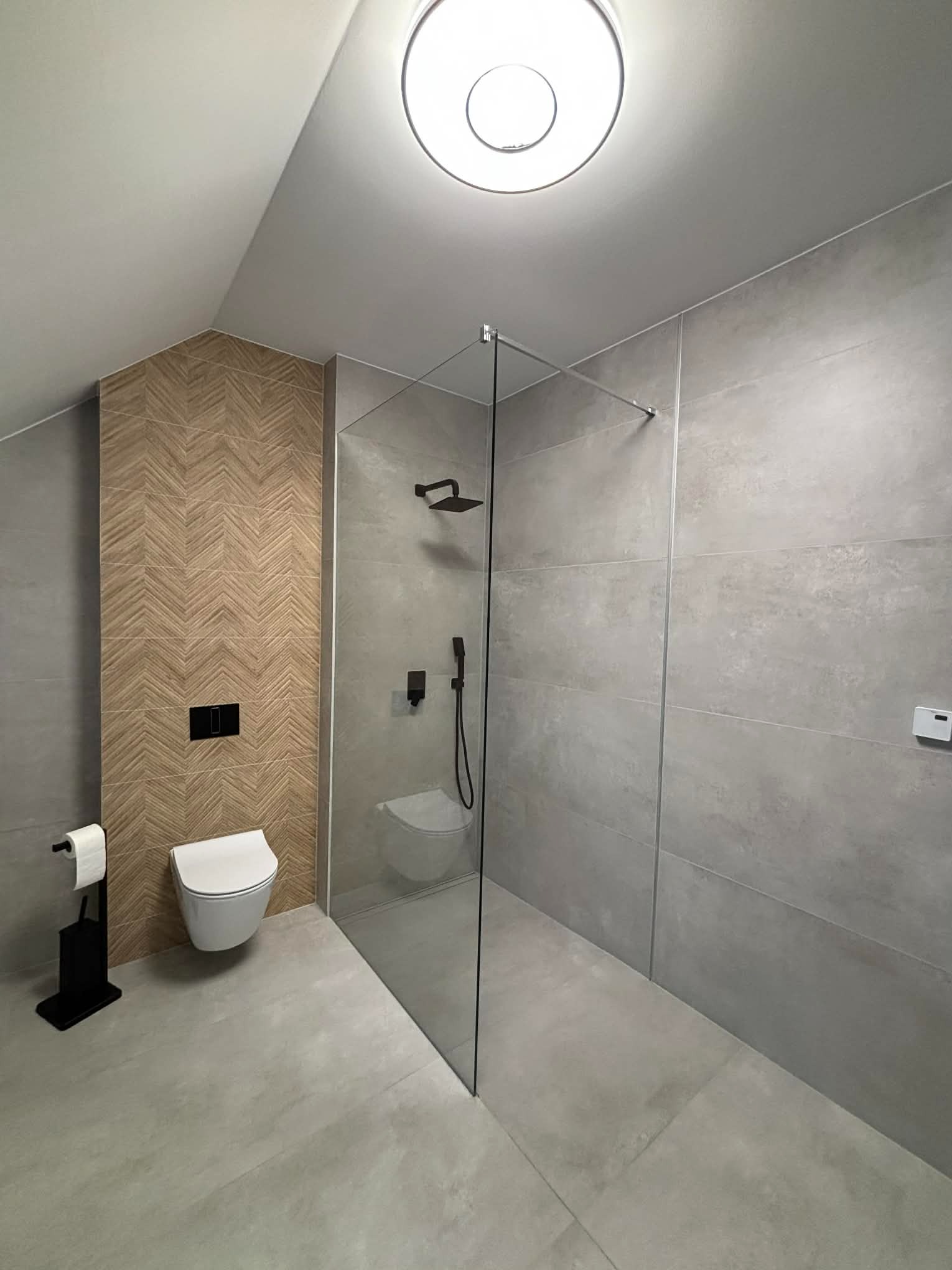Nowoczesna łazienka na poddaszu z kabiną prysznicową typu walk-in, szarą glazurą i akcentem drewnopodobnych płytek ułożonych w jodełkę. Minimalistyczny design.
