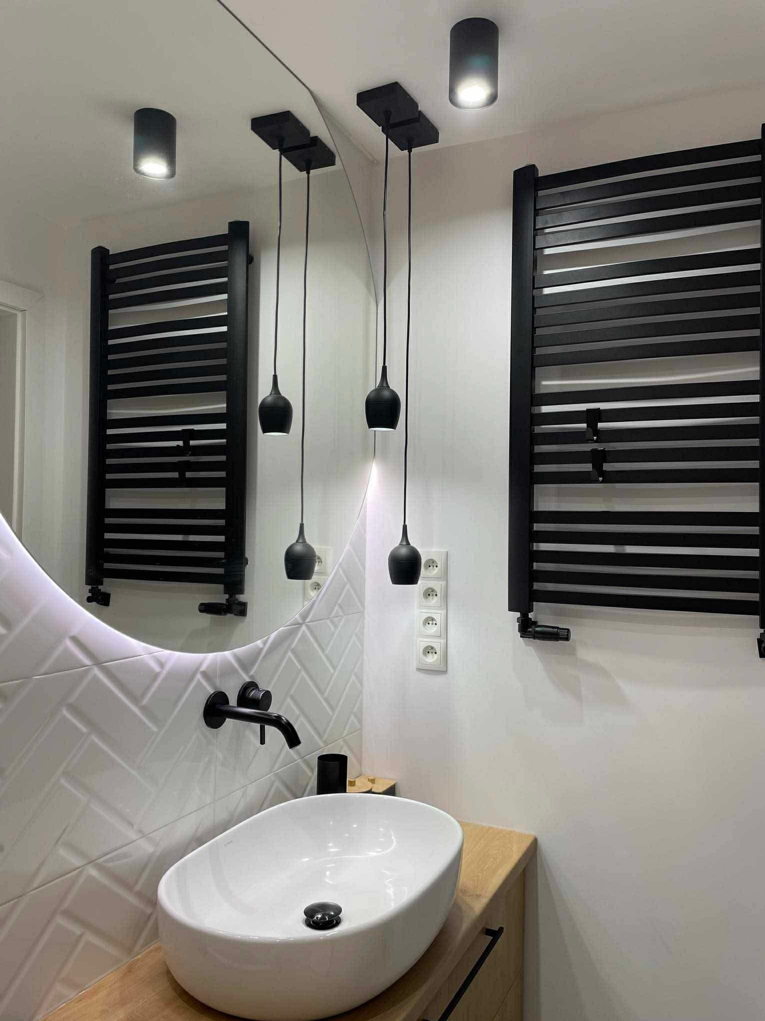 Nowoczesna łazienka z owalną umywalką, czarną armaturą i grzejnikiem drabinkowym. Lustro z podświetleniem LED i wiszące lampy dodają elegancji.