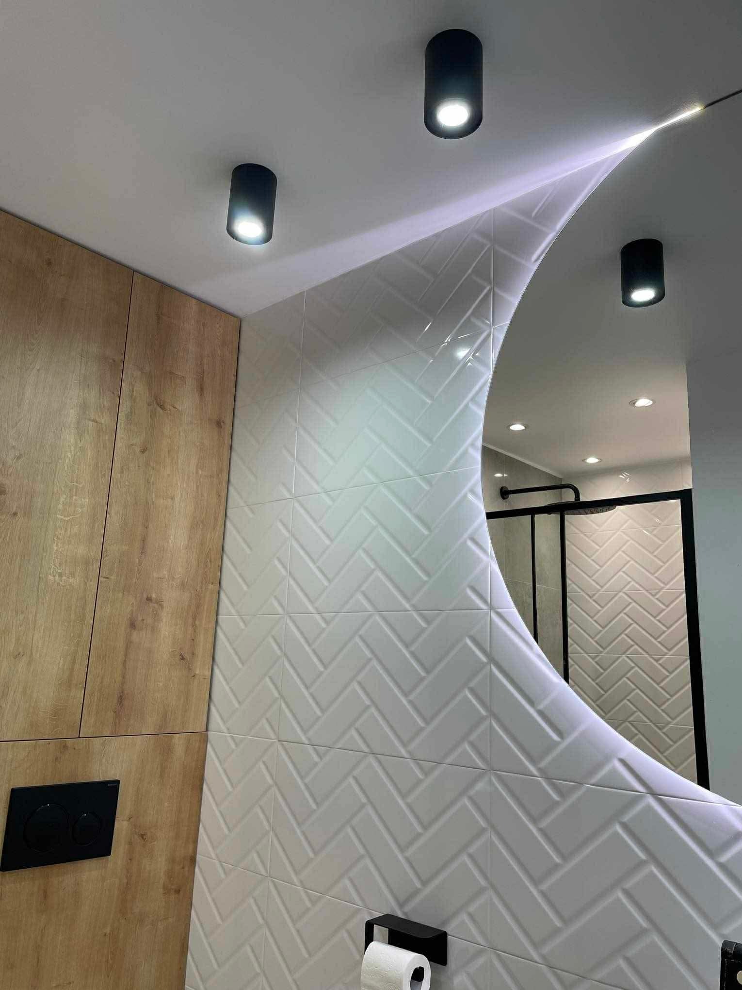 Nowoczesna łazienka po remoncie: białe płytki ułożone w jodełkę, okrągłe lustro z podświetleniem i drewniane szafki. Widoczne czarne, punktowe lampy sufitowe.