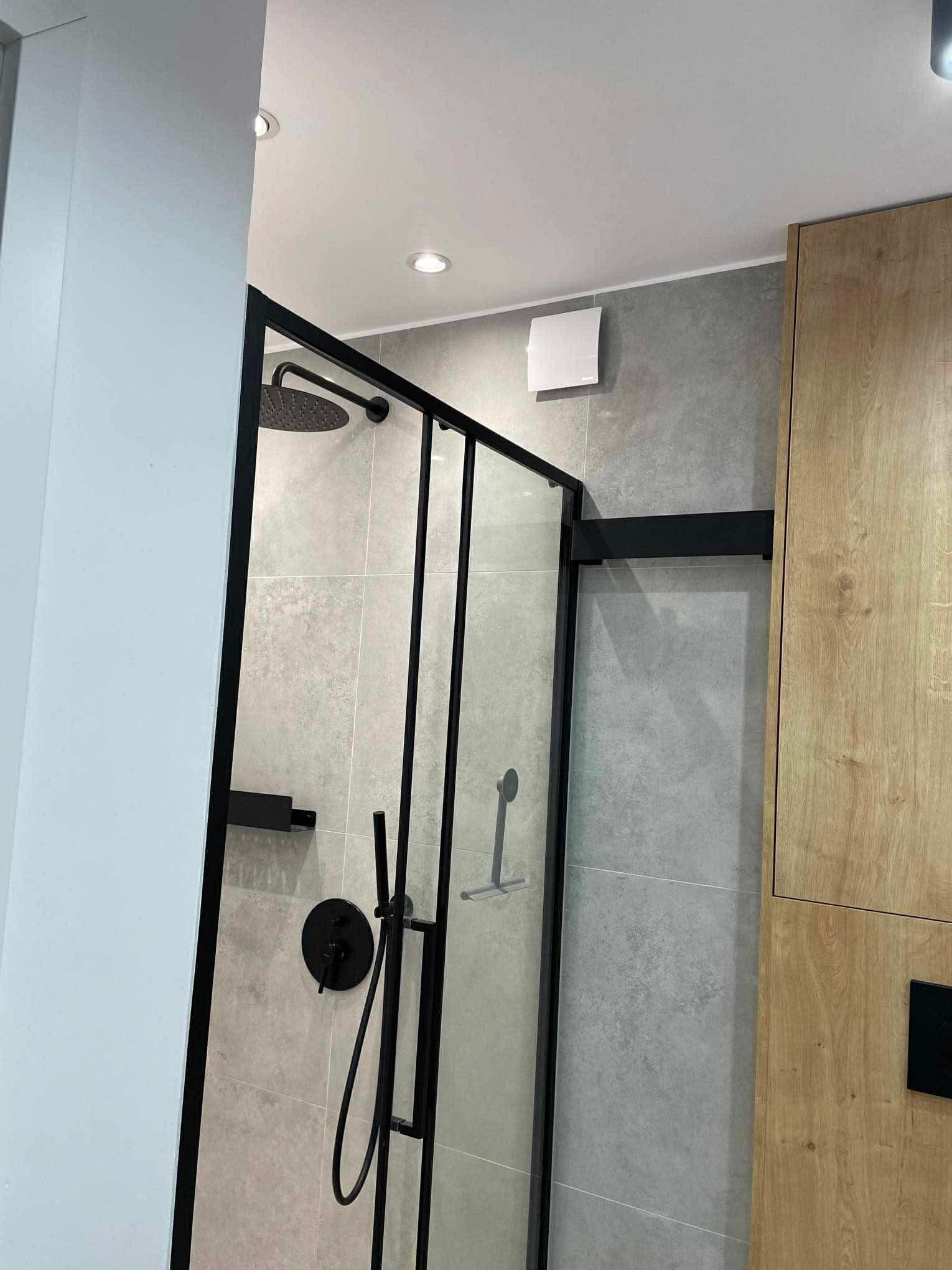 Nowoczesna kabina prysznicowa z czarnymi profilami i deszczownicą, szare płytki na ścianach, drewniana szafka łazienkowa. Minimalistyczny design, oświetlenie punktowe.