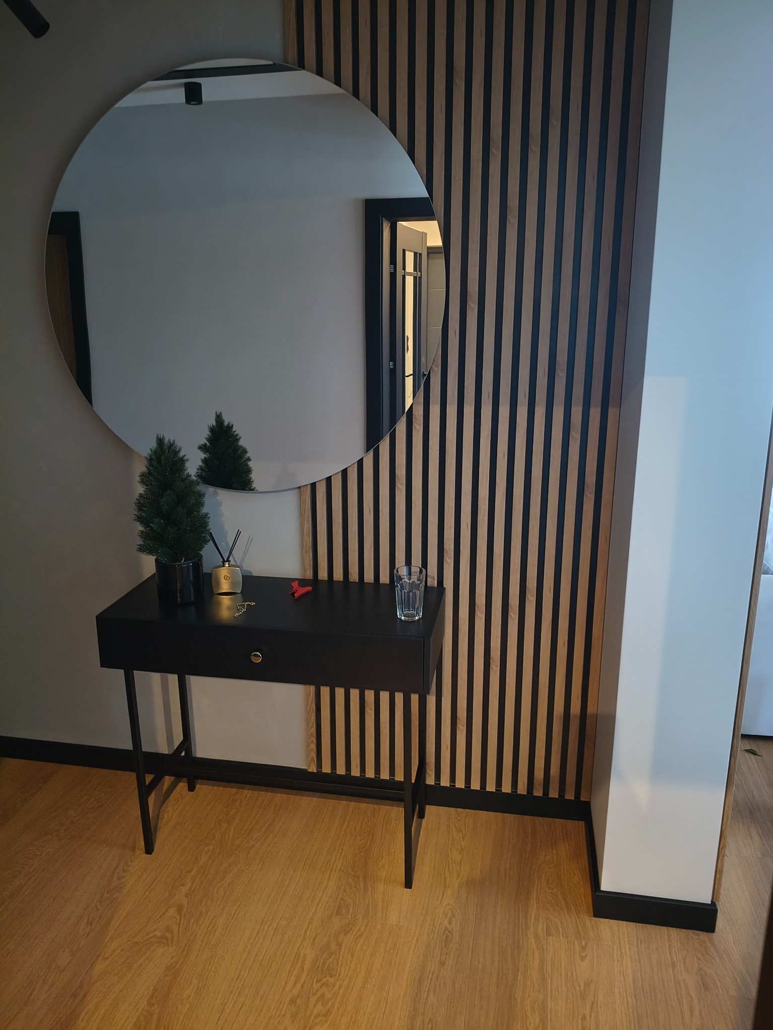 Nowoczesny przedpokój z okrągłym lustrem, konsolą i lamelami na ścianie. Minimalistyczny design z akcentami dekoracyjnymi i ciepłym oświetleniem.