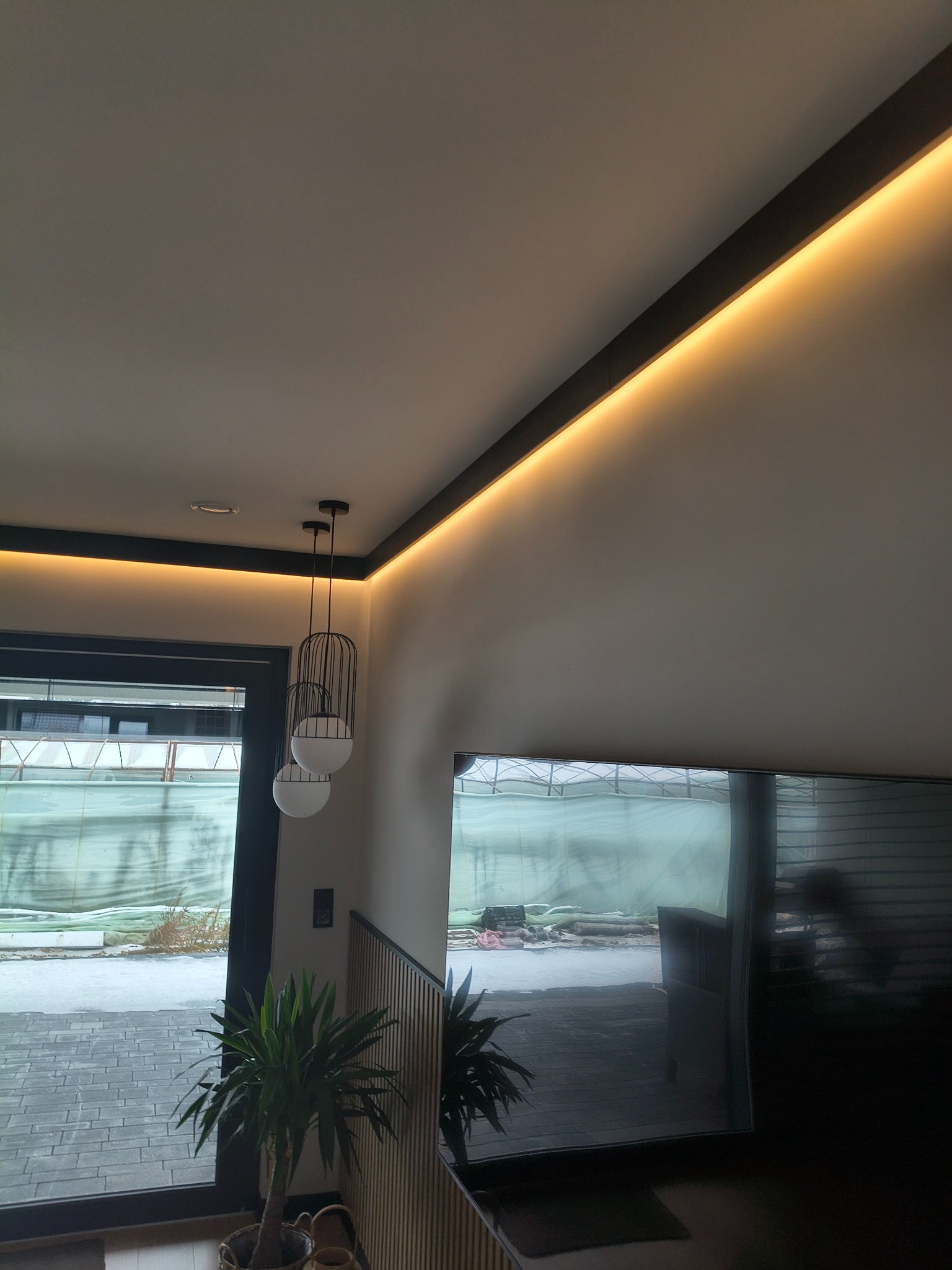 Nowoczesne wnętrze z oświetleniem LED wzdłuż sufitu, wiszące lampy kuliste i widok na zewnątrz przez okno. Minimalistyczny design z roślinami doniczkowymi.