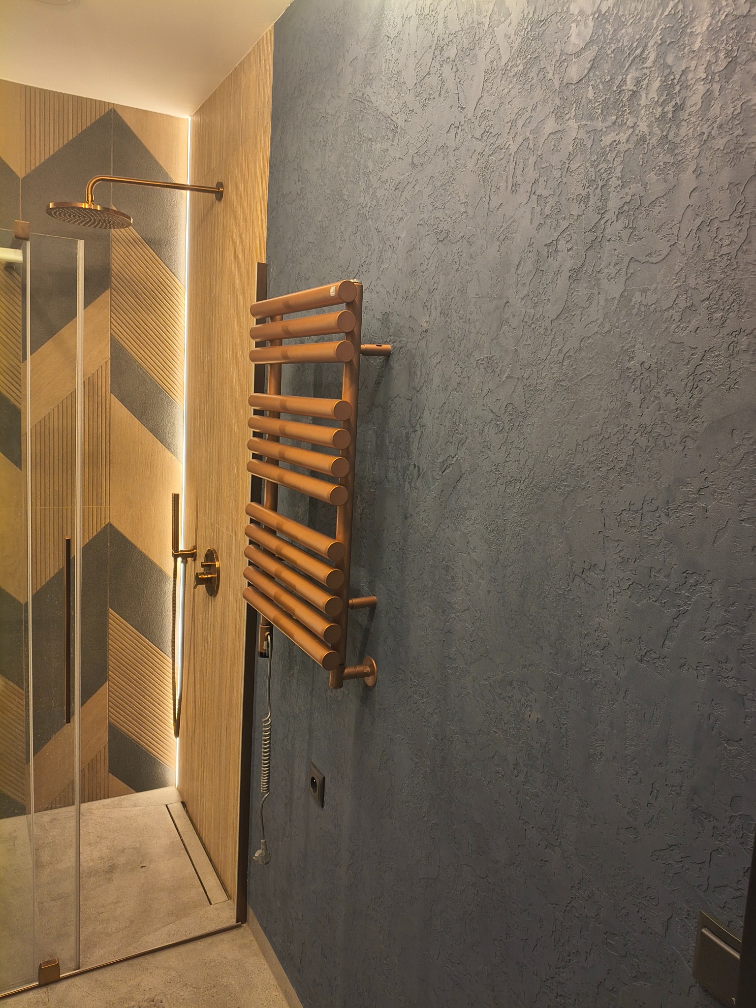 Nowoczesna łazienka z kabiną prysznicową, dekoracyjną ścianą w odcieniach szarości i miedzianym grzejnikiem drabinkowym. Geometryczne wzory na ścianie prysznica.