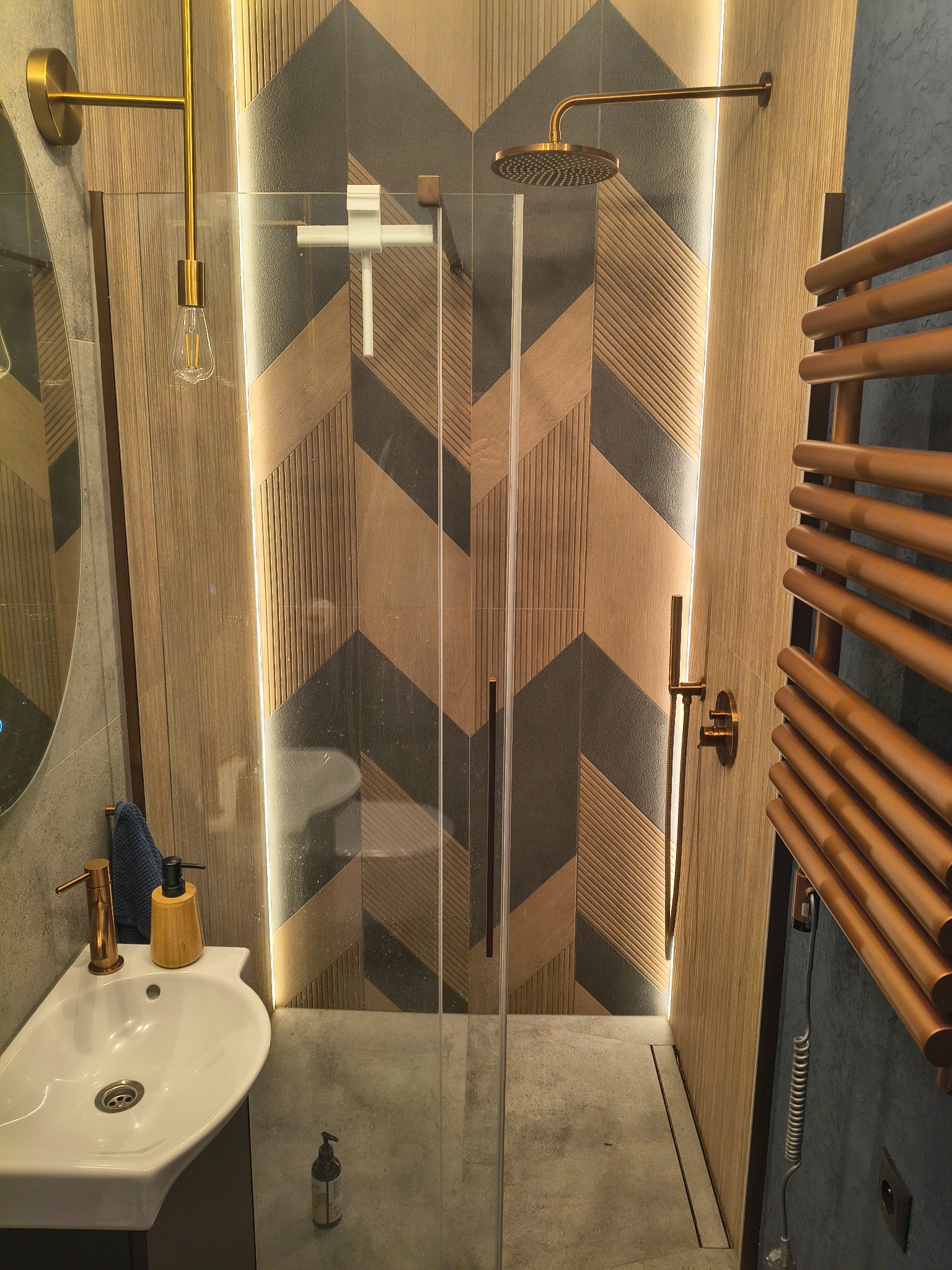 Nowoczesna łazienka z kabiną prysznicową typu walk-in, geometrycznymi płytkami i złotymi akcentami: armatura, oświetlenie i grzejnik drabinkowy. Minimalistyczny design.