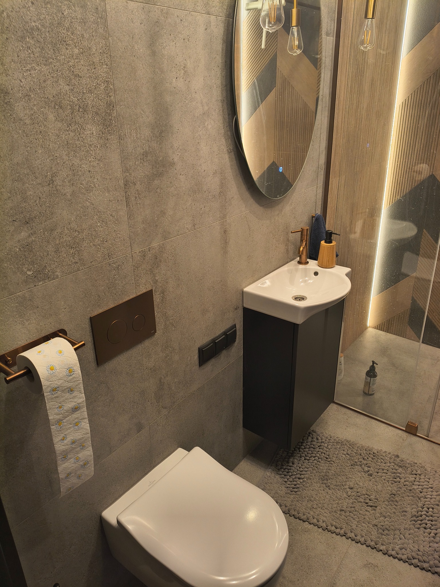 Nowoczesna łazienka w szarościach z owalnym lustrem podświetlanym LED, wiszącą toaletą i umywalką. Armatura w kolorze miedzi. Widoczny fragment kabiny prysznicowej.