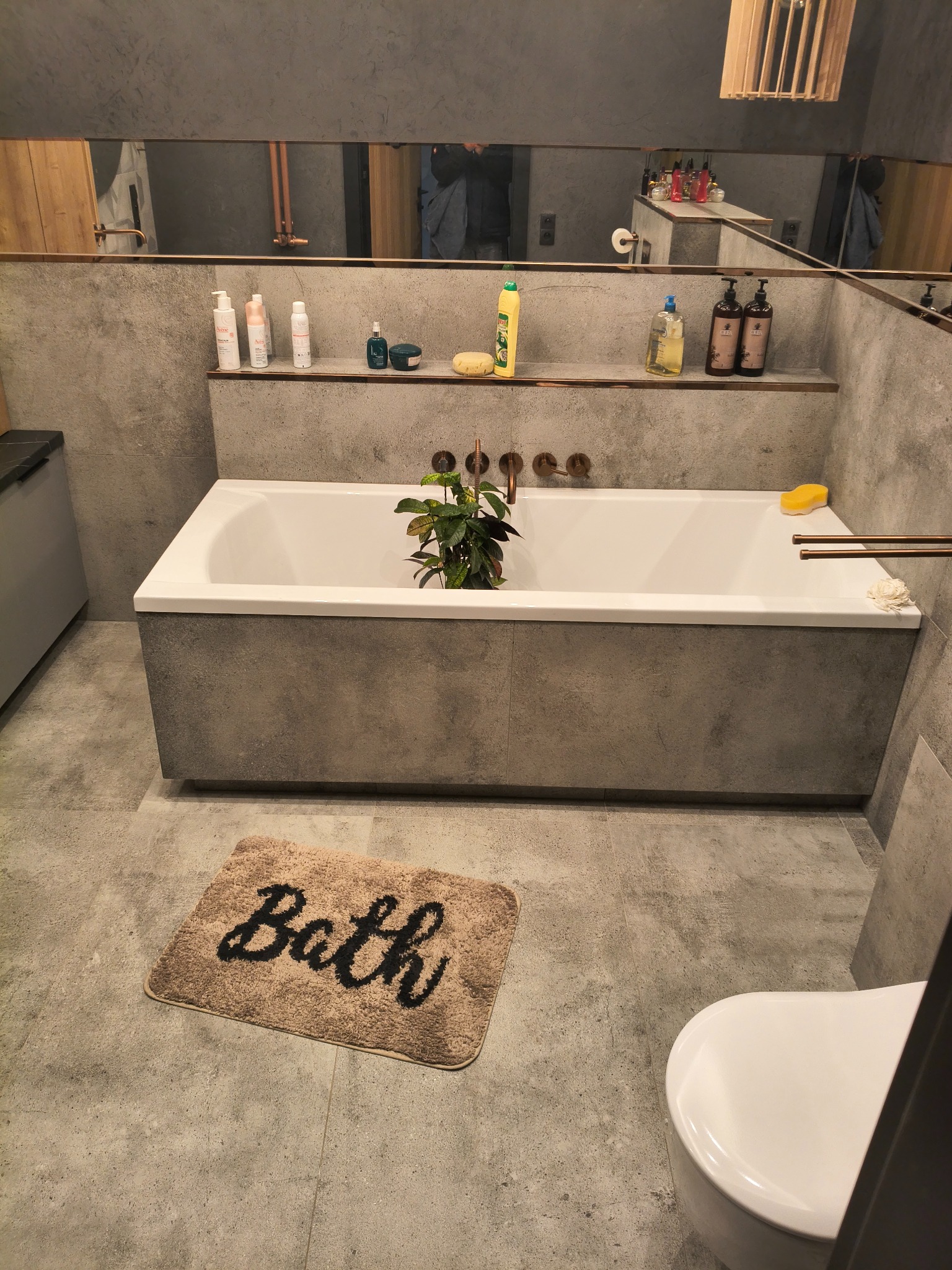 Nowoczesna łazienka z wanną obudowaną szarym kamieniem, armaturą w kolorze miedzi i lustrem na całej szerokości ściany. Na podłodze leży dywanik z napisem 'Bath'.