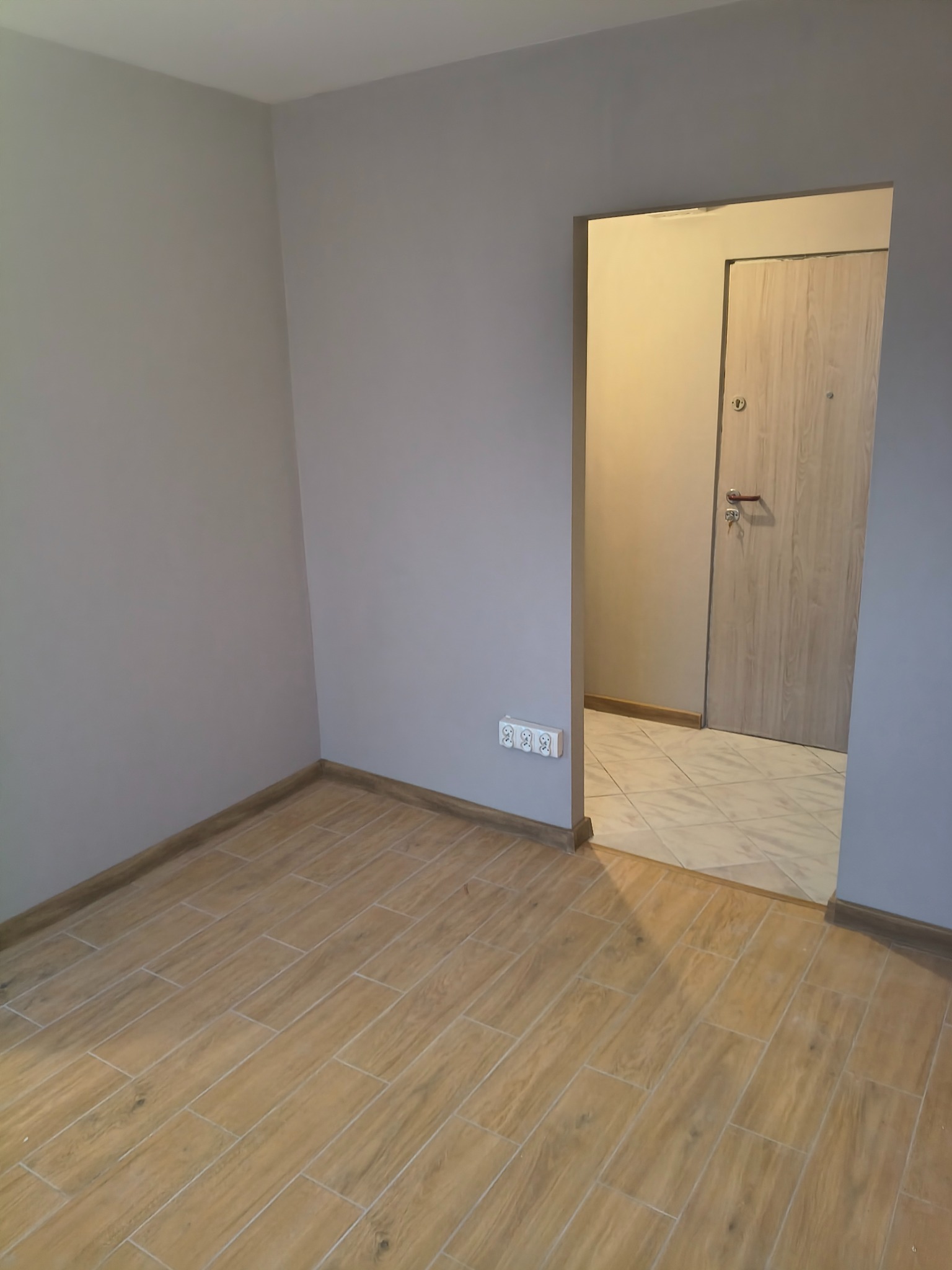 Jasne panele podłogowe imitujące drewno w nowym pokoju z szarą ścianą i widokiem na przedpokój z beżową podłogą i drzwiami.