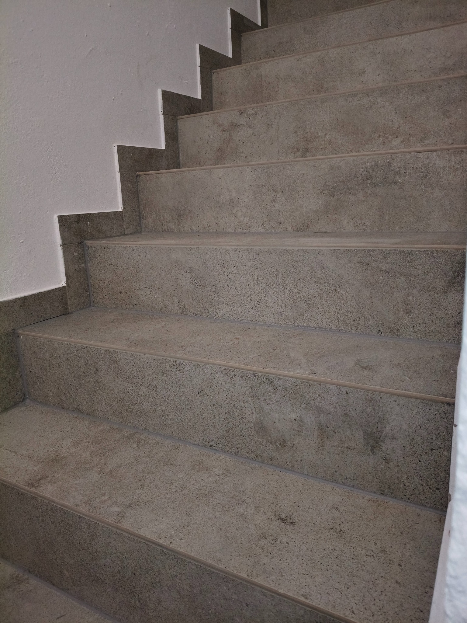 Schody wykończone szarą, kamienną okładziną, widok z dołu. Minimalistyczny design, jasne ściany, precyzyjne wykończenie krawędzi. Detal architektoniczny.