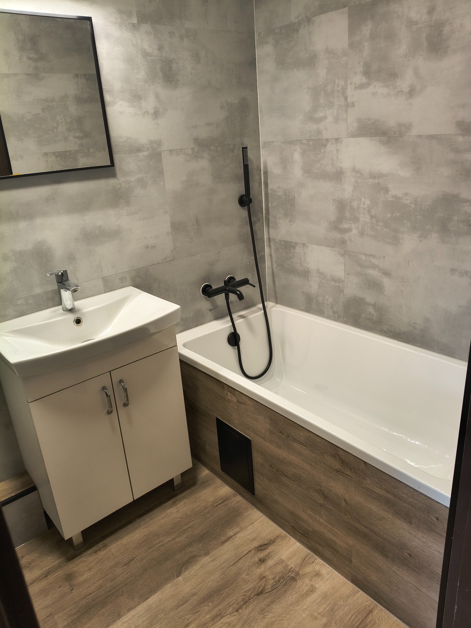 Wykończona łazienka z wanną obudowaną drewnopodobną płytką, umywalką z szafką i szarą płytką na ścianach. Czarna armatura dodaje nowoczesnego charakteru.