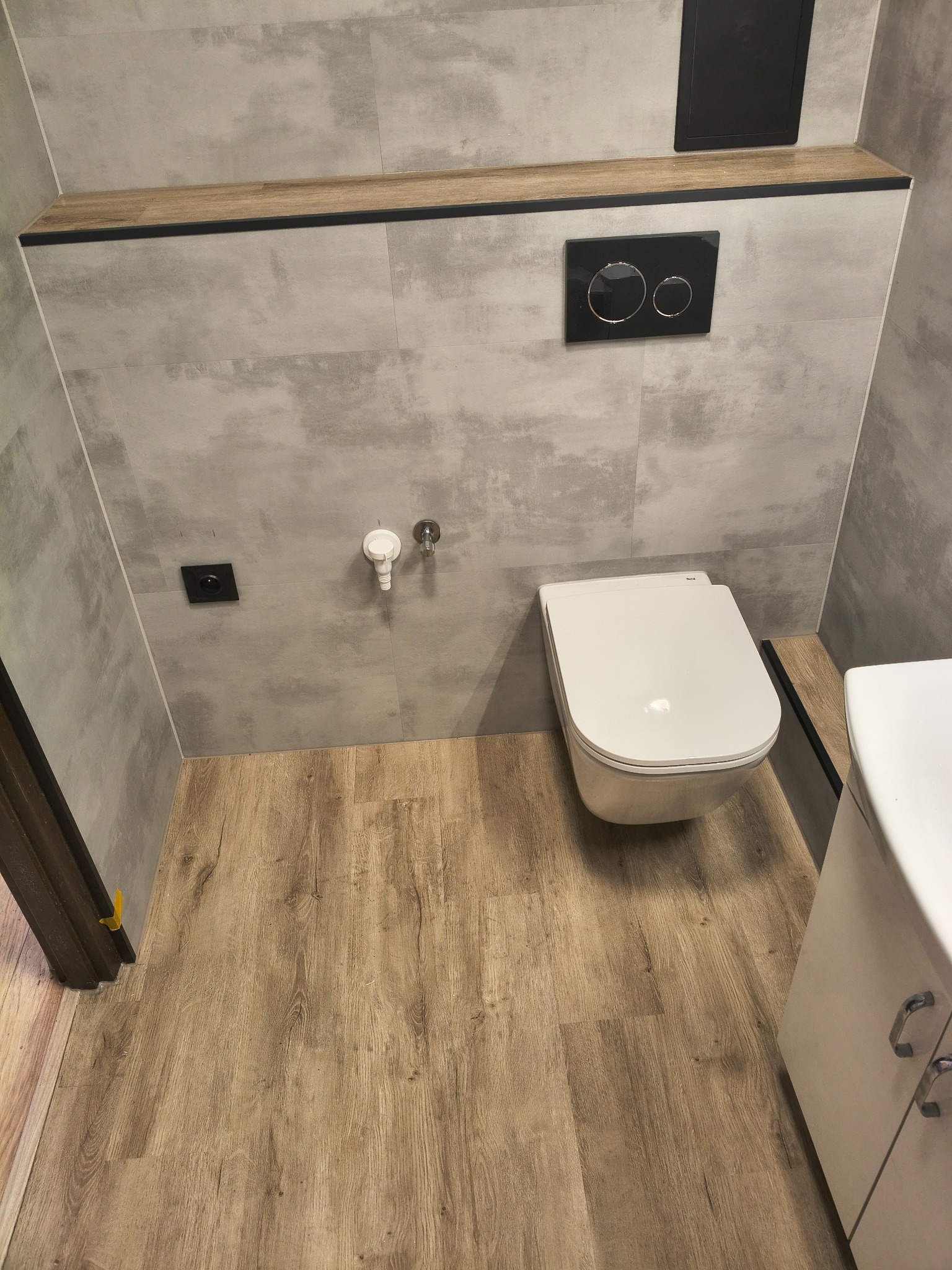 Wykończona łazienka z podwieszaną toaletą, szarą płytką imitującą beton i panelami podłogowymi. Widoczna zabudowa geberit i szafka umywalkowa.