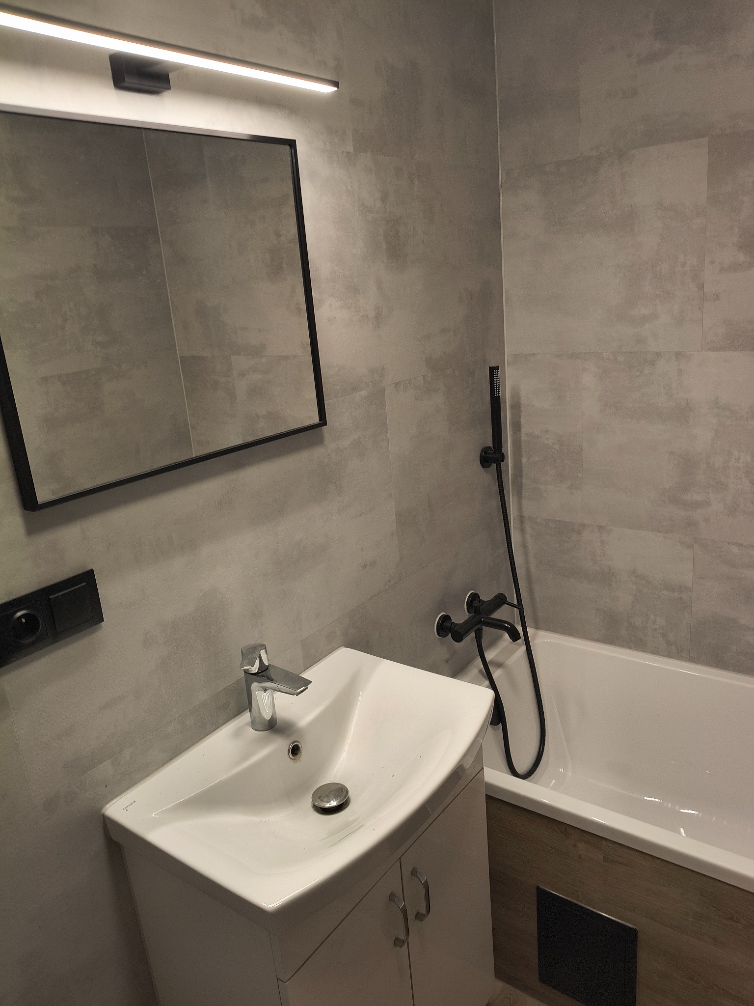 Nowoczesna łazienka po remoncie: biała umywalka z szafką, czarna armatura, lustro z oświetleniem LED i szare płytki imitujące beton. Minimalistyczny design.