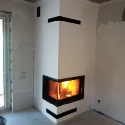 Nowoczesny narożny kominek z czarnymi akcentami w surowym wnętrzu, widoczne płomienie w palenisku, ściany w stanie deweloperskim z widocznymi śladami prac wykończeniowych.