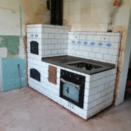 Kuchenny piec kaflowy z piekarnikiem i fajerkami, wykończony białymi kaflami z niebieskim wzorem, w surowym wnętrzu podczas remontu.