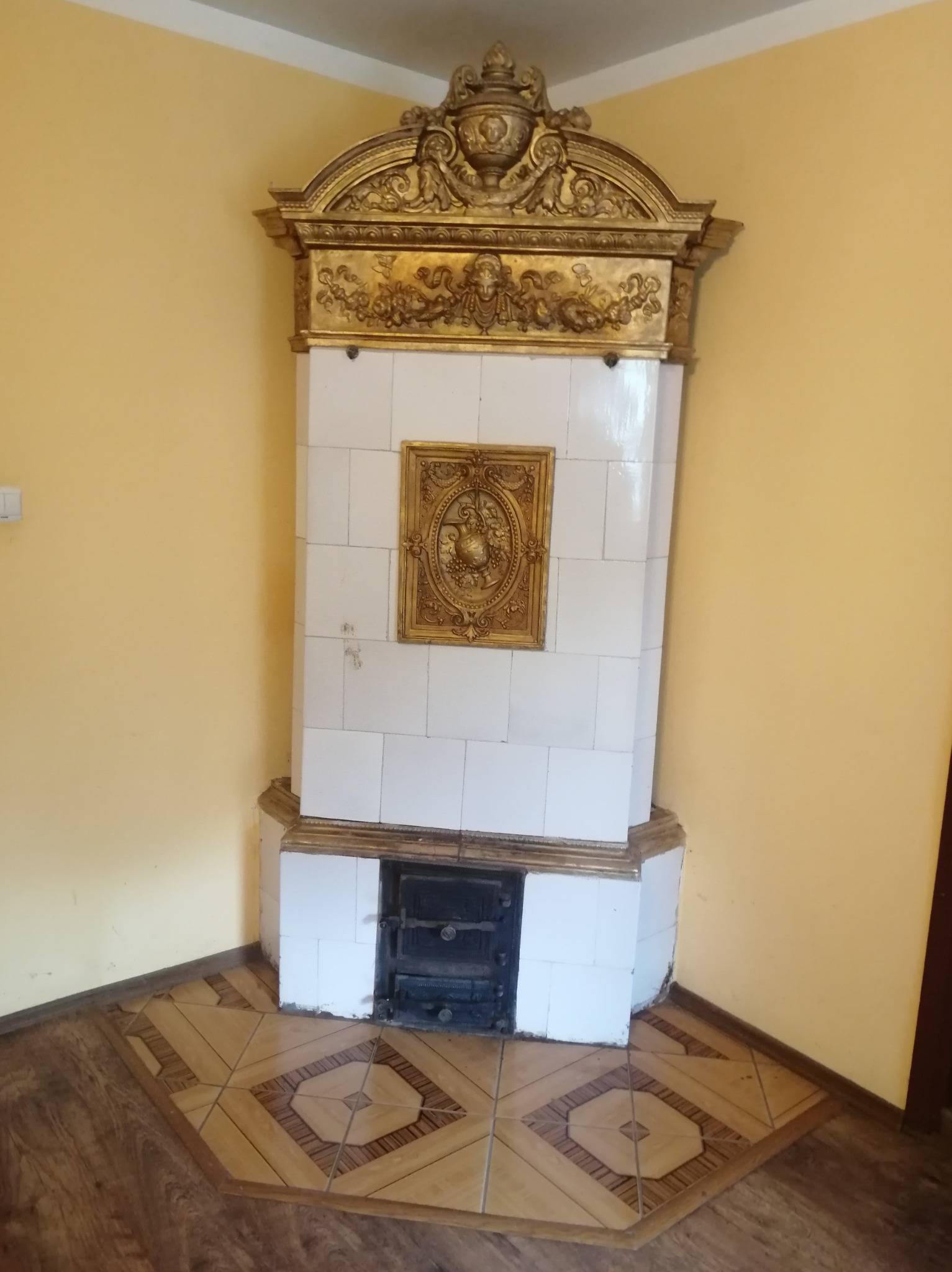 Biały piec kaflowy z dekoracyjną, złoconą obudową w rogu pokoju, z widocznymi drzwiczkami paleniska i fragmentem podłogi z ceramicznymi płytkami.