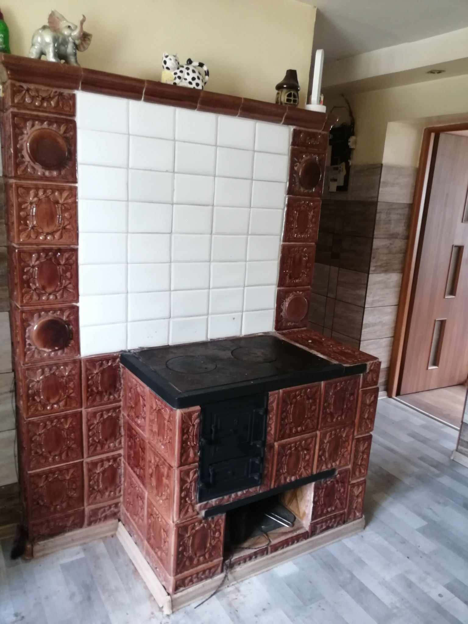 Rustykalny piec kaflowy z czarnym blatem i drzwiczkami, zdobiony brązowymi kaflami z motywem roślinnym, zwieńczony białymi kaflami i ozdobnymi figurkami.