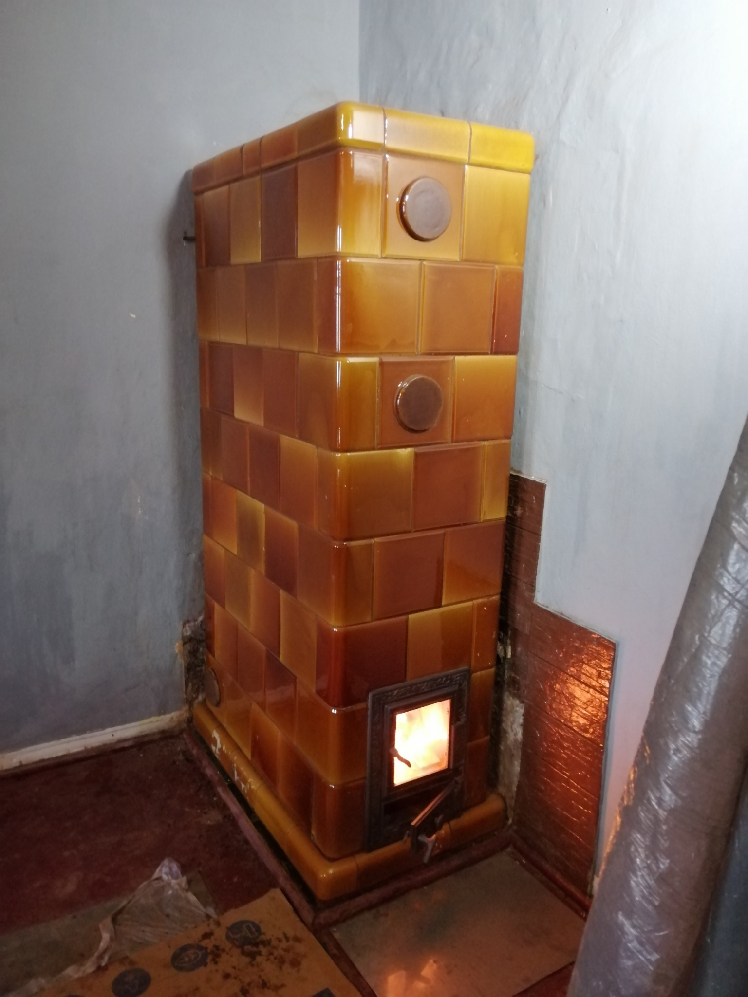 Kaflowy piec kominkowy w odcieniach brązu i żółci, z widocznym płomieniem w palenisku, ustawiony w rogu pomieszczenia z szarą ścianą.