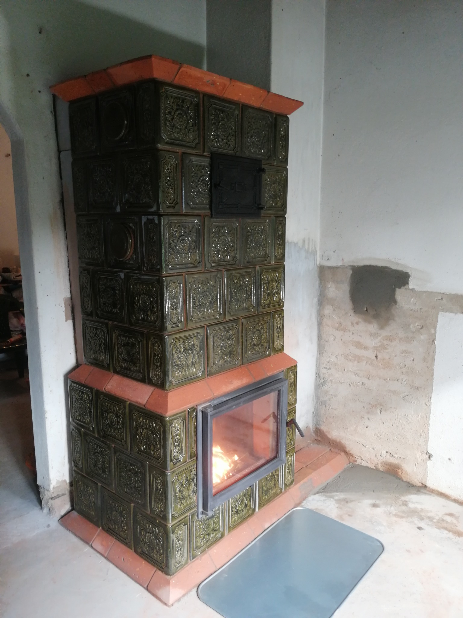 Piec kaflowy z paleniskiem, obudowany ciemnozielonymi kaflami z ornamentami, obramowany czerwoną cegłą. W palenisku płonie ogień. Widoczne ślady prac murarskich na ścianie.