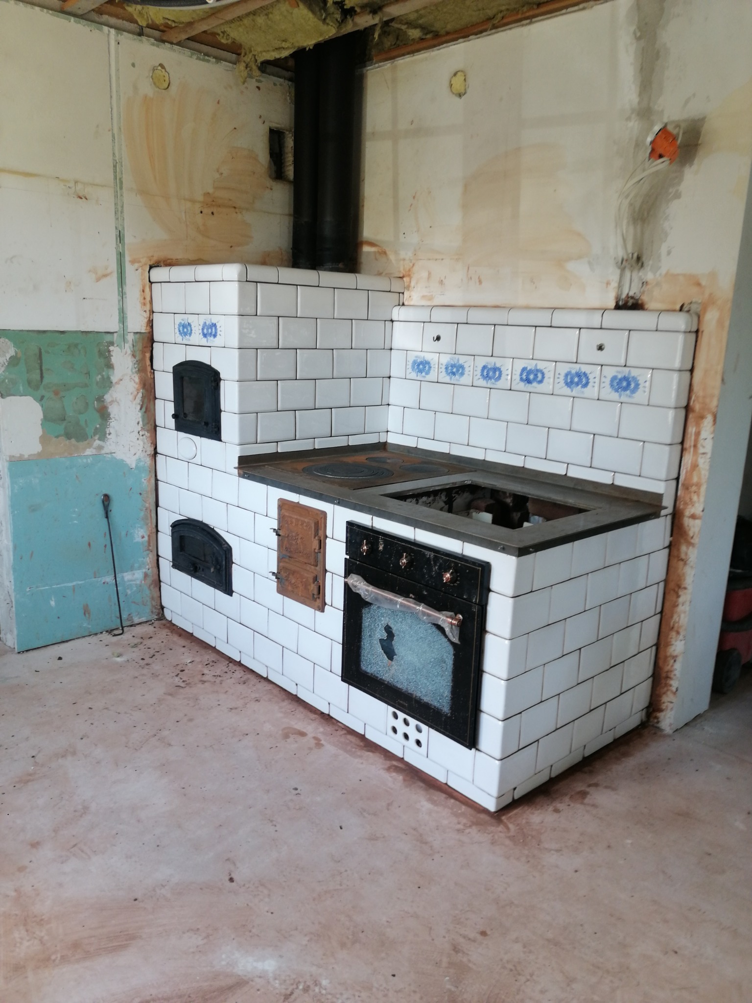 Kuchenny piec kaflowy z piekarnikiem i fajerkami, wykończony białymi kaflami z niebieskim wzorem, w surowym wnętrzu podczas remontu.