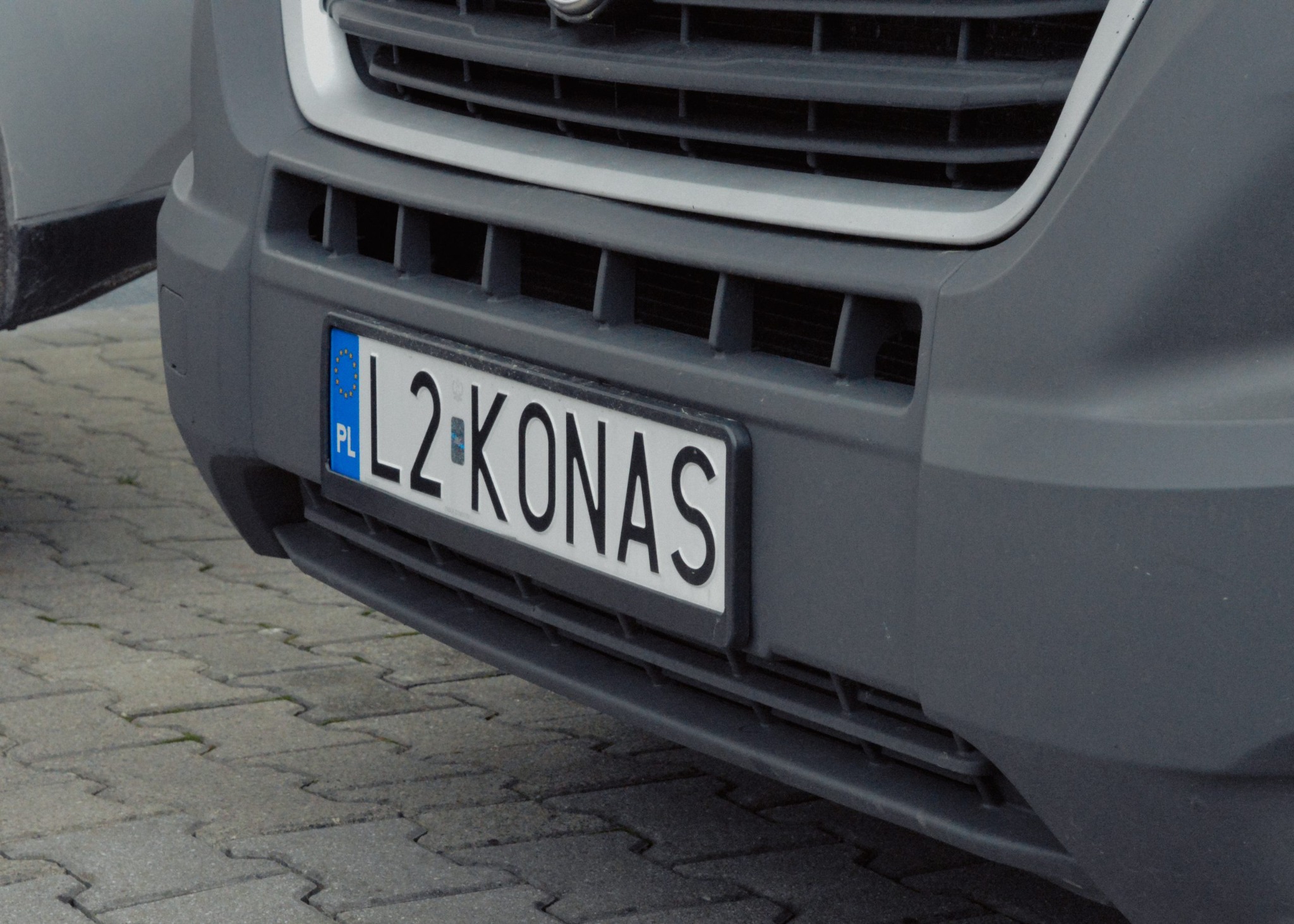 Szary samochód dostawczy z polską tablicą rejestracyjną L2-KONAS, zaparkowany na kostce brukowej.