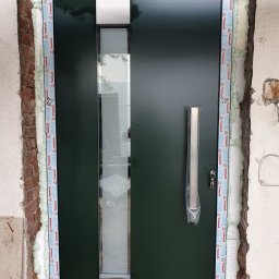 Oknopast Piotr Pasternak - Ciemnozielone drzwi wejściowe z długim, pionowym przeszkleniem i srebrnym uchwytem w folii ochronnej, osadzone w surowym otworze ściennym z widoczną pianką montażową i elementami muru.