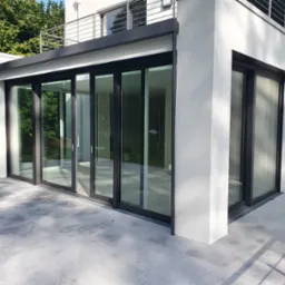 Nowoczesny przeszklony taras z przesuwnymi drzwiami, czarne ramy, szara posadzka z płytek, widok z zewnątrz, balkon na górze.