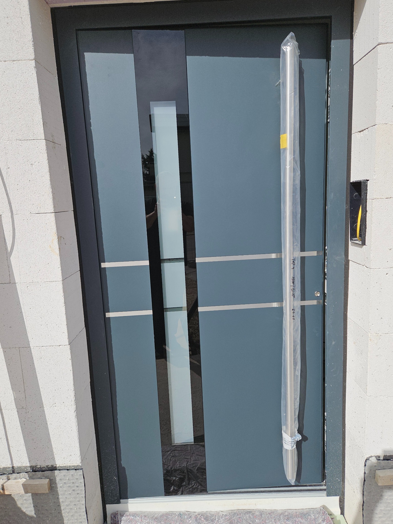 Nowoczesne, szare drzwi wejściowe z przeszkleniami, zamontowane w surowym murze, z uchwytem w folii ochronnej, widoczny domofon po prawej stronie.