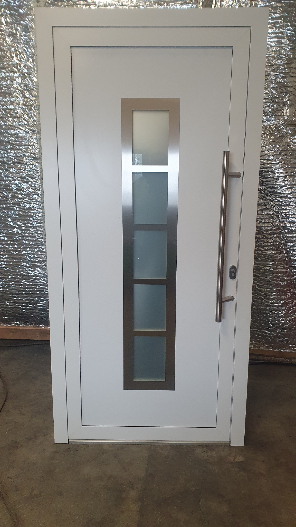 Białe drzwi zewnętrzne z prostokątnym przeszkleniem z czterema matowymi szybami i długim, srebrnym uchwytem, sfotografowane na tle ściany pokrytej folią izolacyjną.