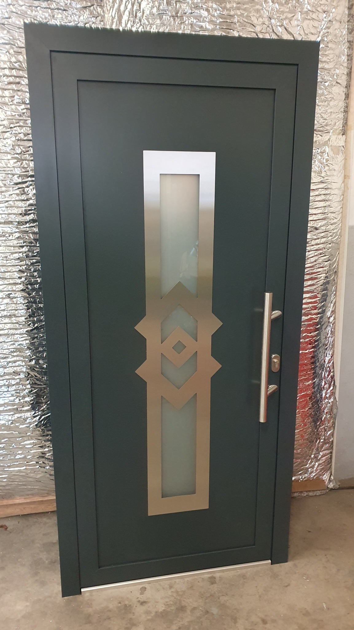 Ciemnozielone drzwi zewnętrzne z podłużnym, matowym przeszkleniem zdobionym geometrycznym wzorem w kolorze złotym i srebrną klamką.